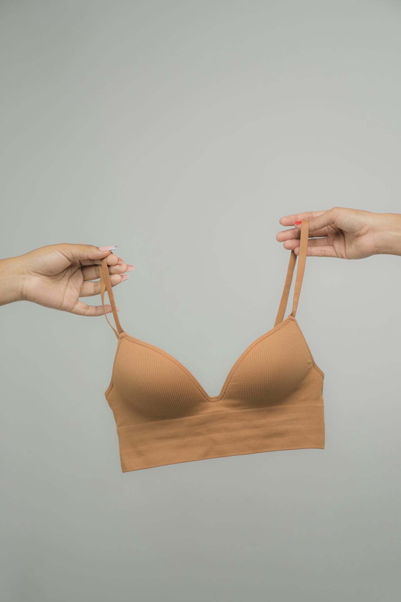 Minimalist close-up of a beige bra held by two hands against a neutral backdrop.